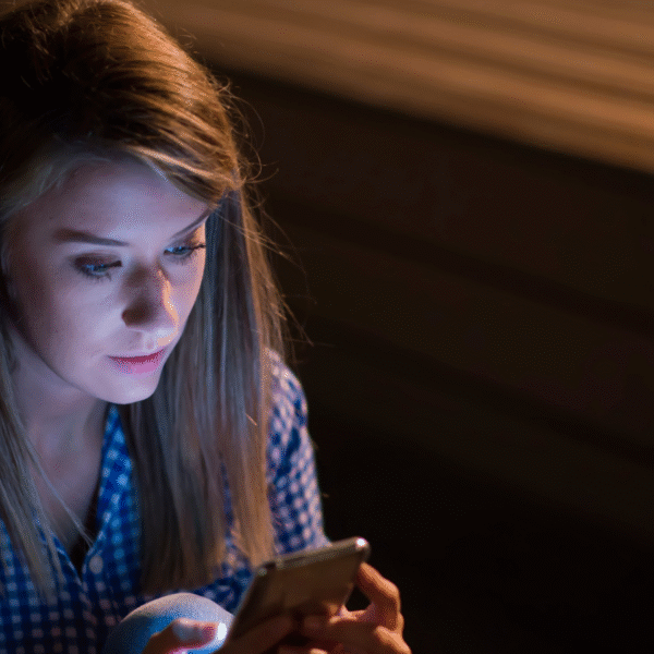 සයිබර් අපරාධ - Young woman sitting alone at night, looking at her smartphone screen with concern, symbolizing the risks of sharing private photos online and the dangers of cyber crime.”