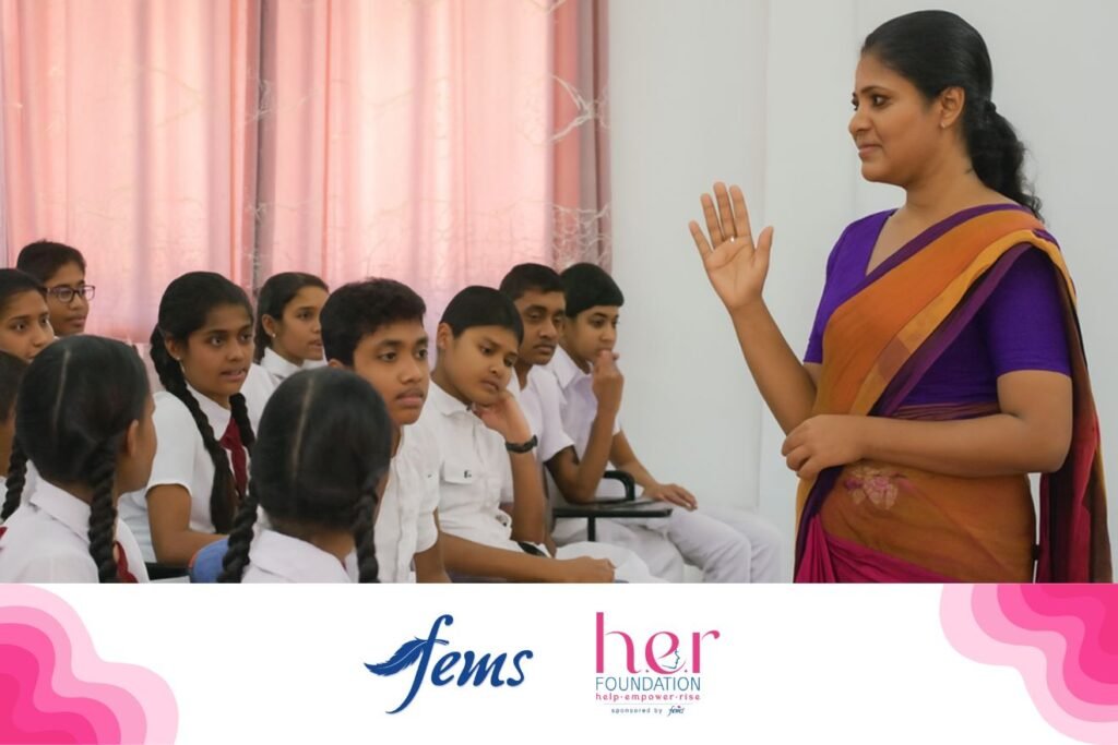 පිරිමි ප්‍රජාව | A female teacher wearing a purple and orange saree speaks to a group of Sri Lankan school students, both boys and girls, during an awareness session. Logos of Fems and the HER Foundation appear at the bottom, highlighting a menstrual health education program.