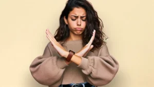 Boundaries | Young woman wearing a beige sweater crossing her arms in an “X” gesture to signal “no”, expressing refusal and setting personal boundaries against a neutral beige background.