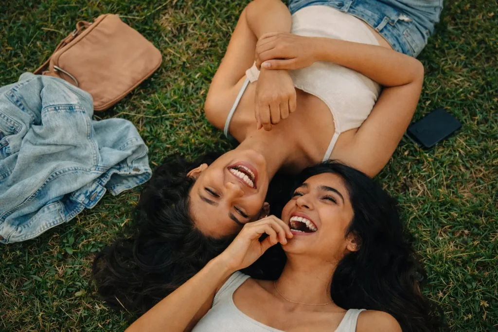 Friendship Audit | Two young Sri Lankan women lying on green grass outdoors, laughing together with their heads close, surrounded by casual items like a bag and jacket, capturing a joyful and natural friendship moment