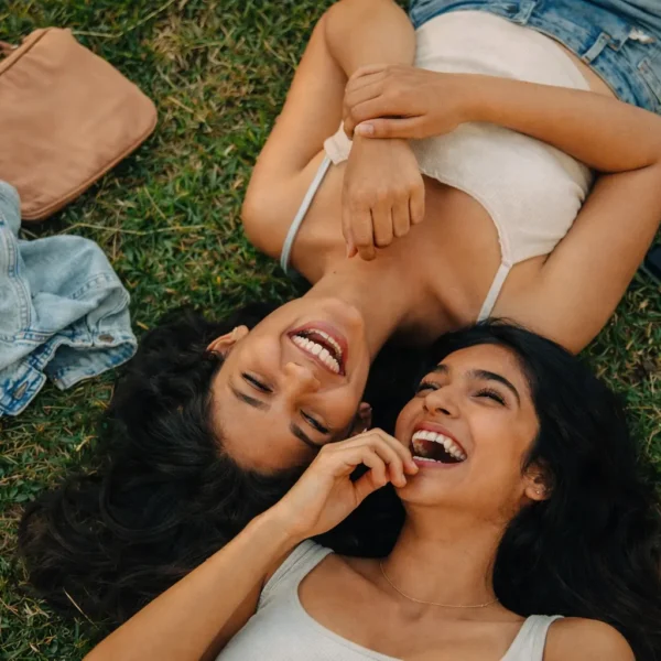 Friendship Audit | Two young Sri Lankan women lying on green grass outdoors, laughing together with their heads close, surrounded by casual items like a bag and jacket, capturing a joyful and natural friendship moment