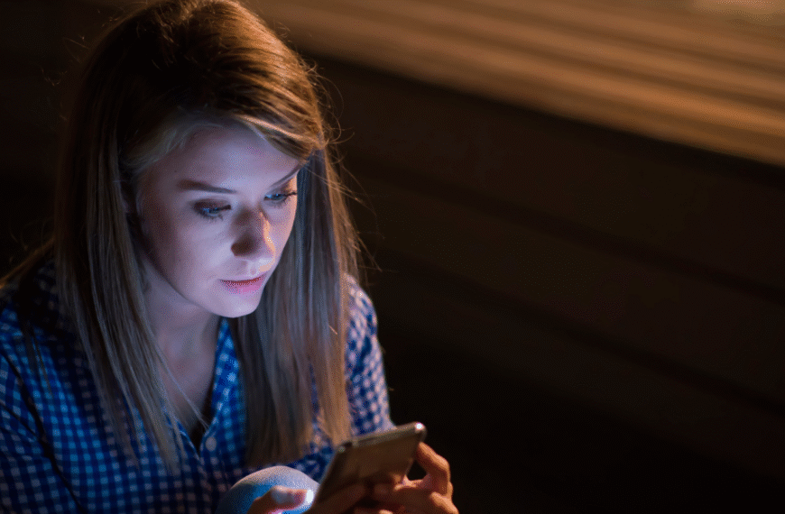 සයිබර් අපරාධ - Young woman sitting alone at night, looking at her smartphone screen with concern, symbolizing the risks of sharing private photos online and the dangers of cyber crime.”