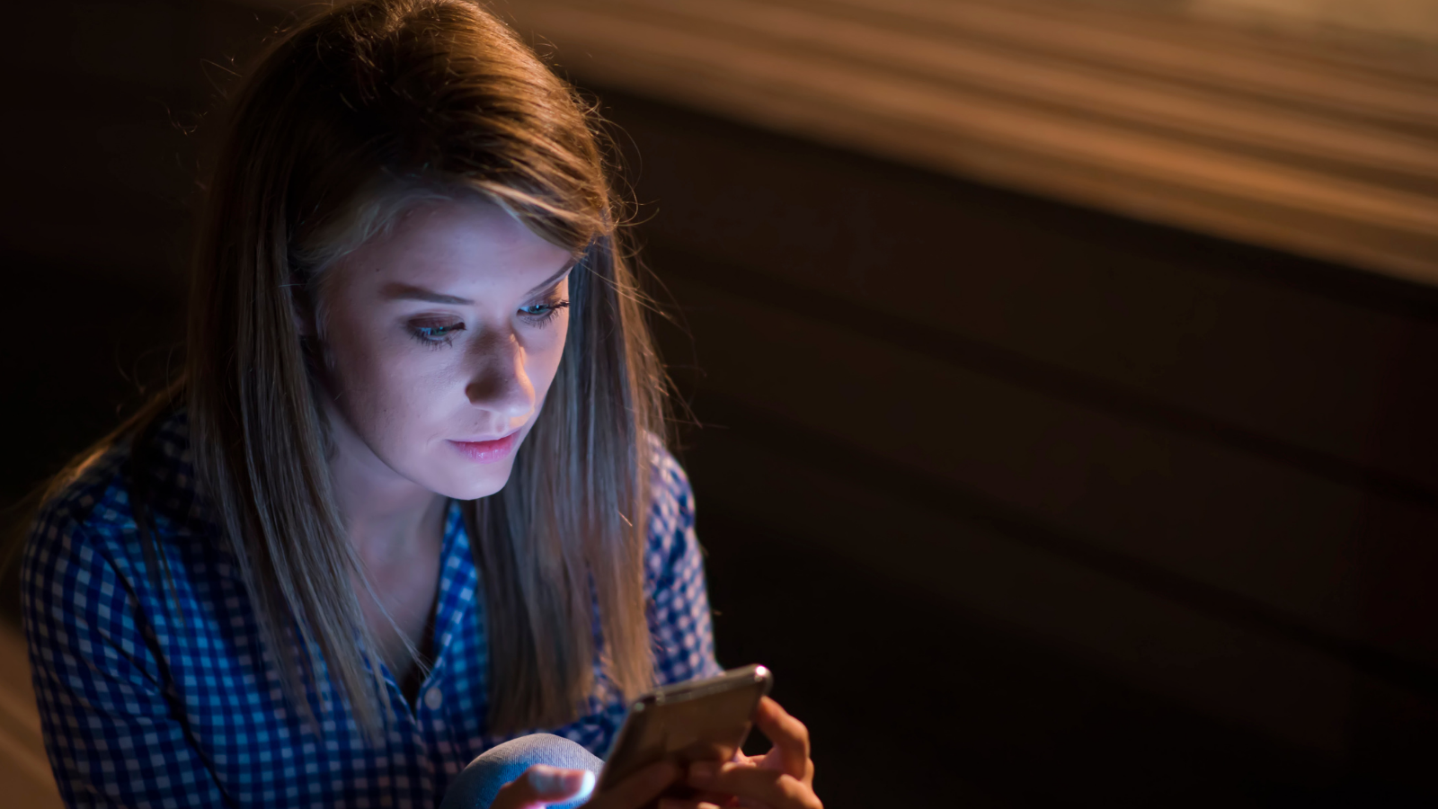සයිබර් අපරාධ - Young woman sitting alone at night, looking at her smartphone screen with concern, symbolizing the risks of sharing private photos online and the dangers of cyber crime.”