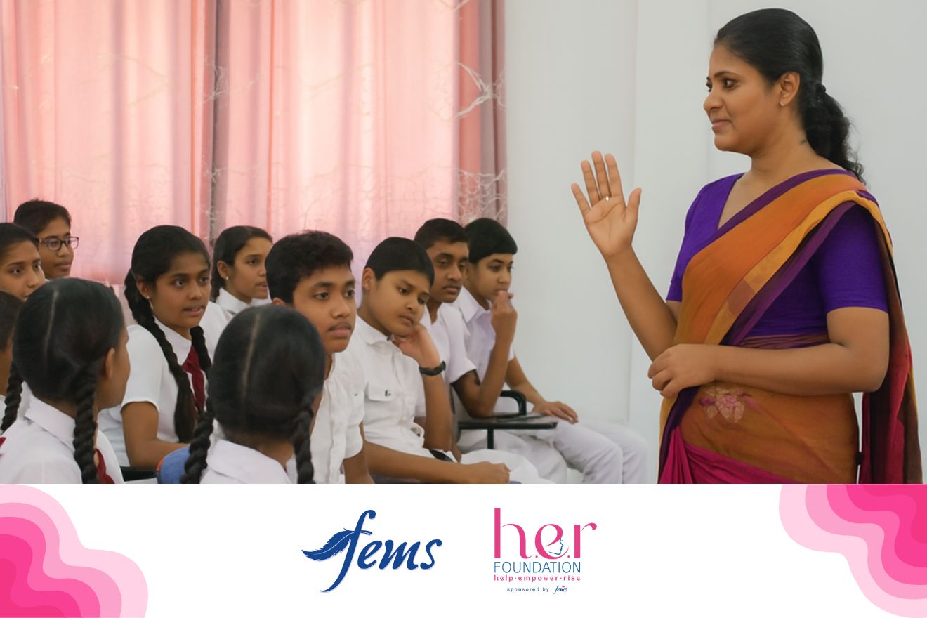 පිරිමි ප්‍රජාව | A female teacher wearing a purple and orange saree speaks to a group of Sri Lankan school students, both boys and girls, during an awareness session. Logos of Fems and the HER Foundation appear at the bottom, highlighting a menstrual health education program.