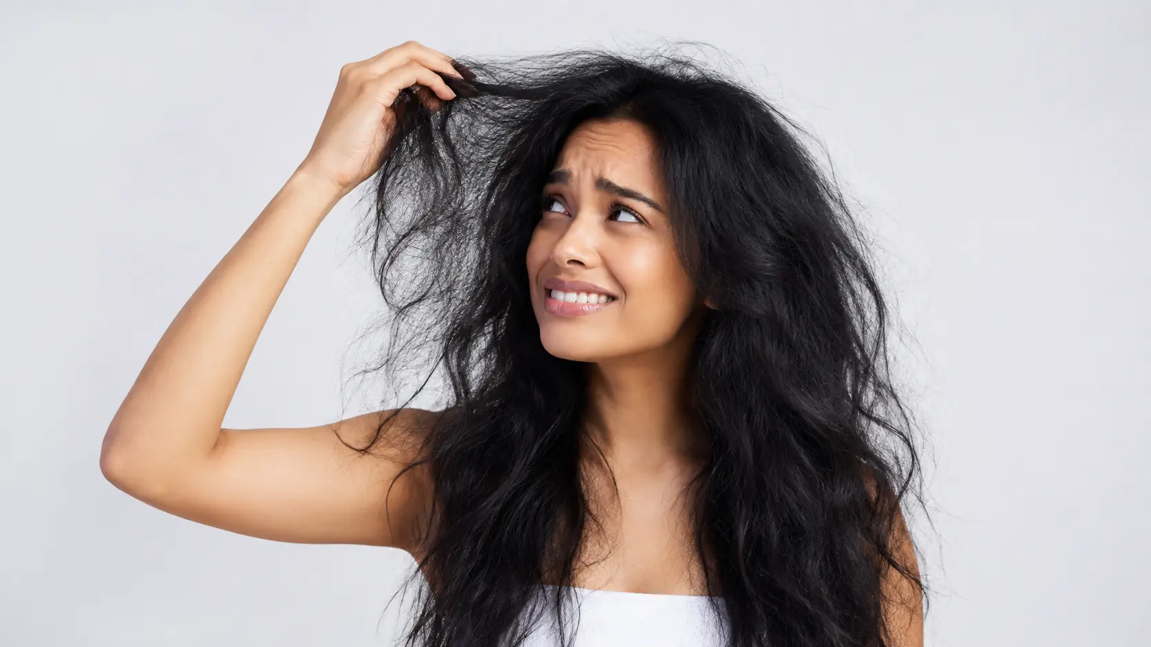 Humidity | A young woman with long dark hair looking concerned as she holds a frizzy, tangled section of her hair, highlighting the effects of humidity, dryness, and hair damage against a clean studio background.