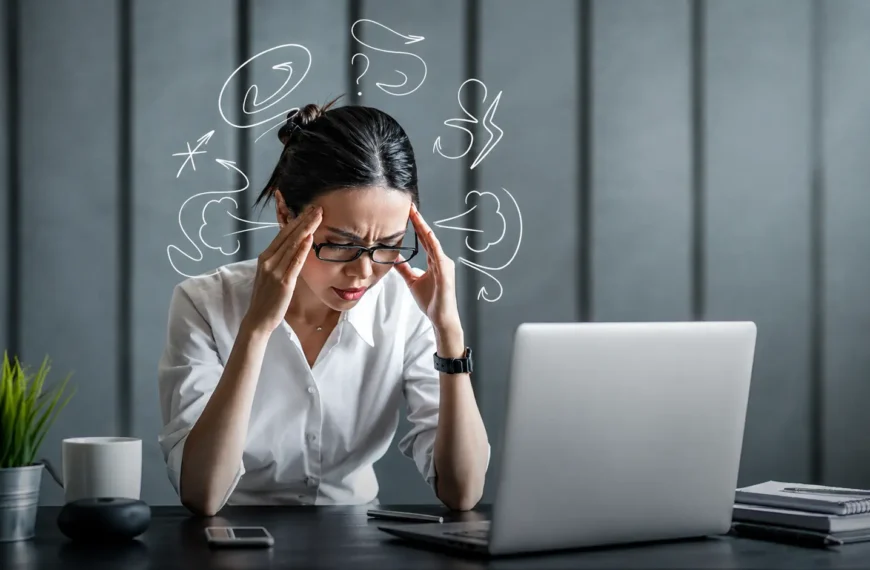 Perfect | Stressed professional woman holding her temples while working at a desk, illustrating perfectionism, overthinking, mental pressure, and workplace stress.