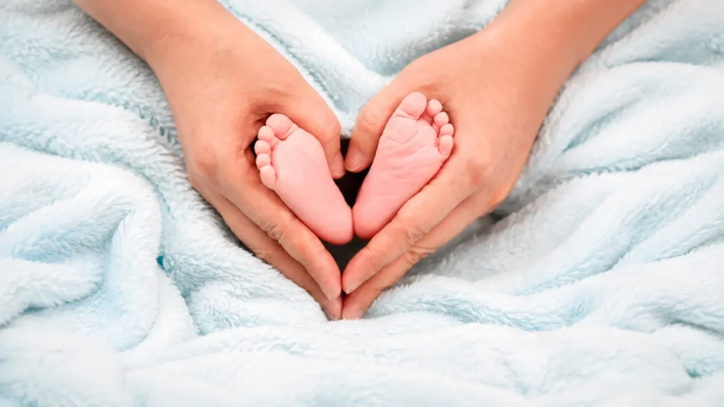 Pregnancy | Adult hands gently holding newborn baby feet on a soft blue blanket.