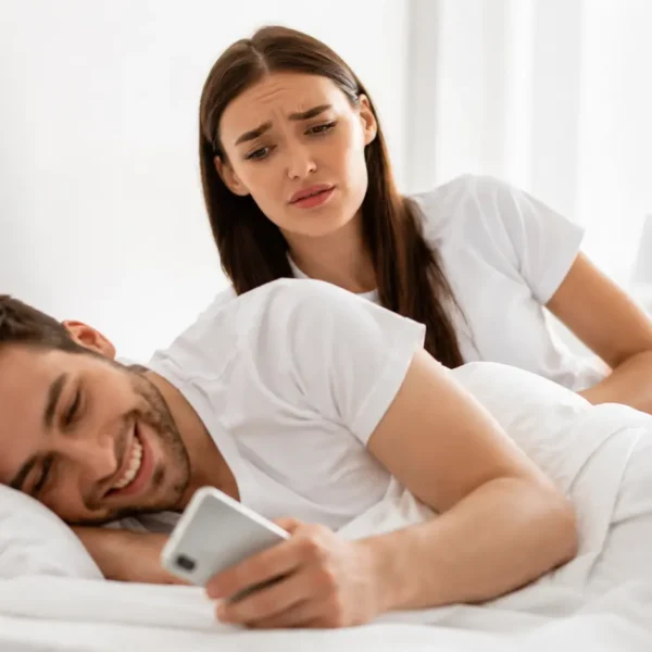 Micro-Cheating | Worried woman looking at her partner using a phone in bed.