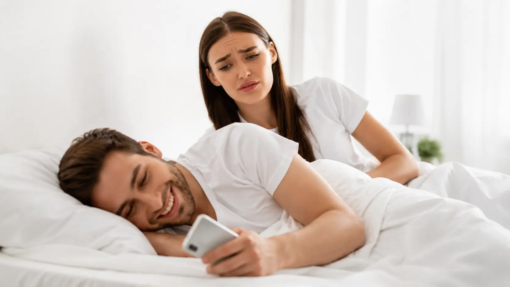 Micro-Cheating | Worried woman looking at her partner using a phone in bed.